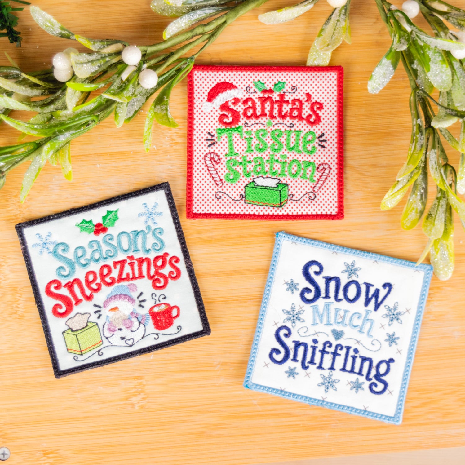 Three embroidered Christmas-themed square patches displayed on a wooden surface with decorative evergreen branches. The patches read 'Santa's Tissue Station,' 'Seasons Sneezings,' and 'Snow Much Sniffling.
