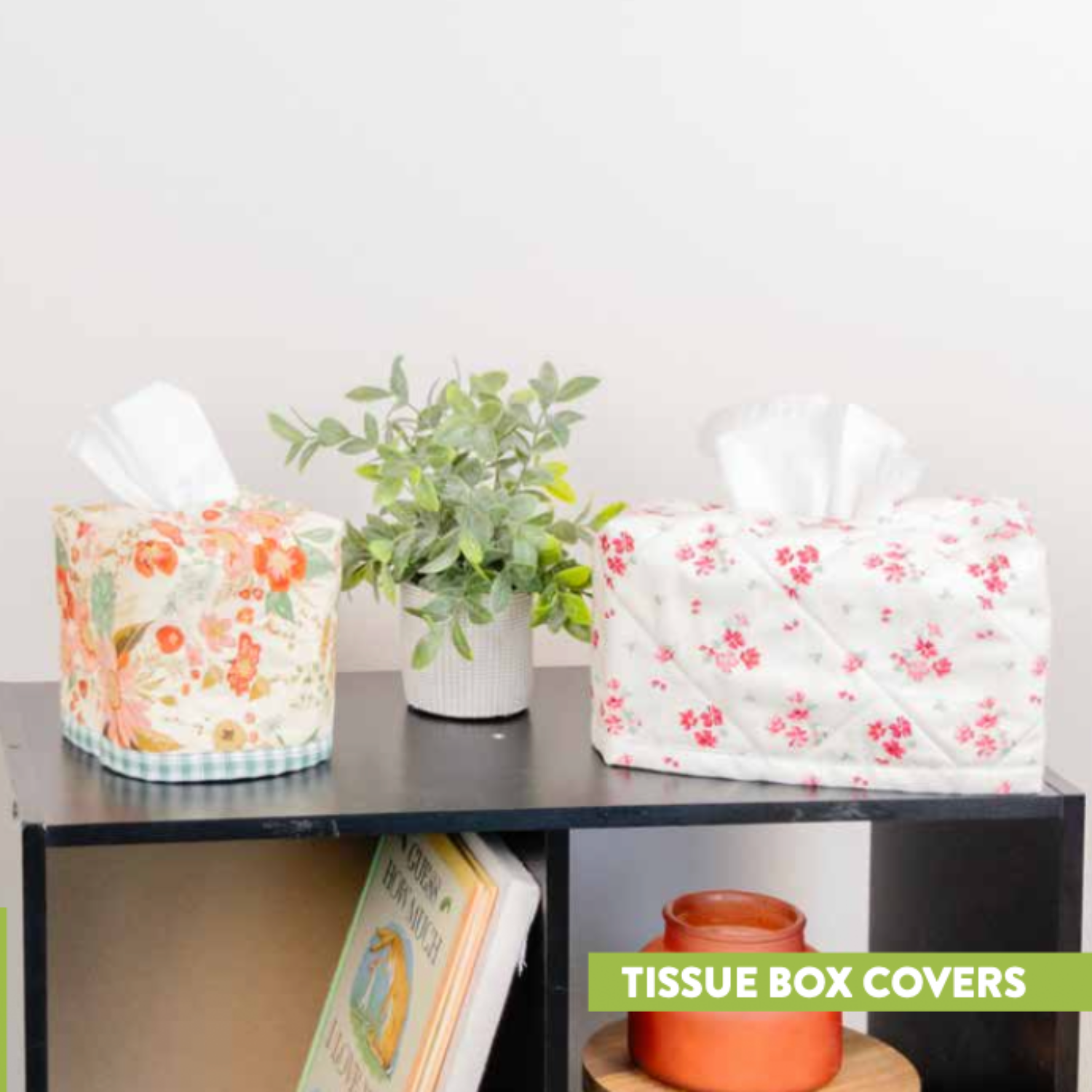Floral tissue box covers on a shelf with a plant and books.