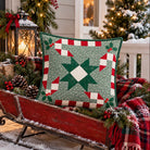 A square quilted throw pillow with a green and red Christmas star pattern sits on a red wooden sleigh decorated with pine branches, pinecones, and a red plaid blanket, set against a festive holiday porch background.