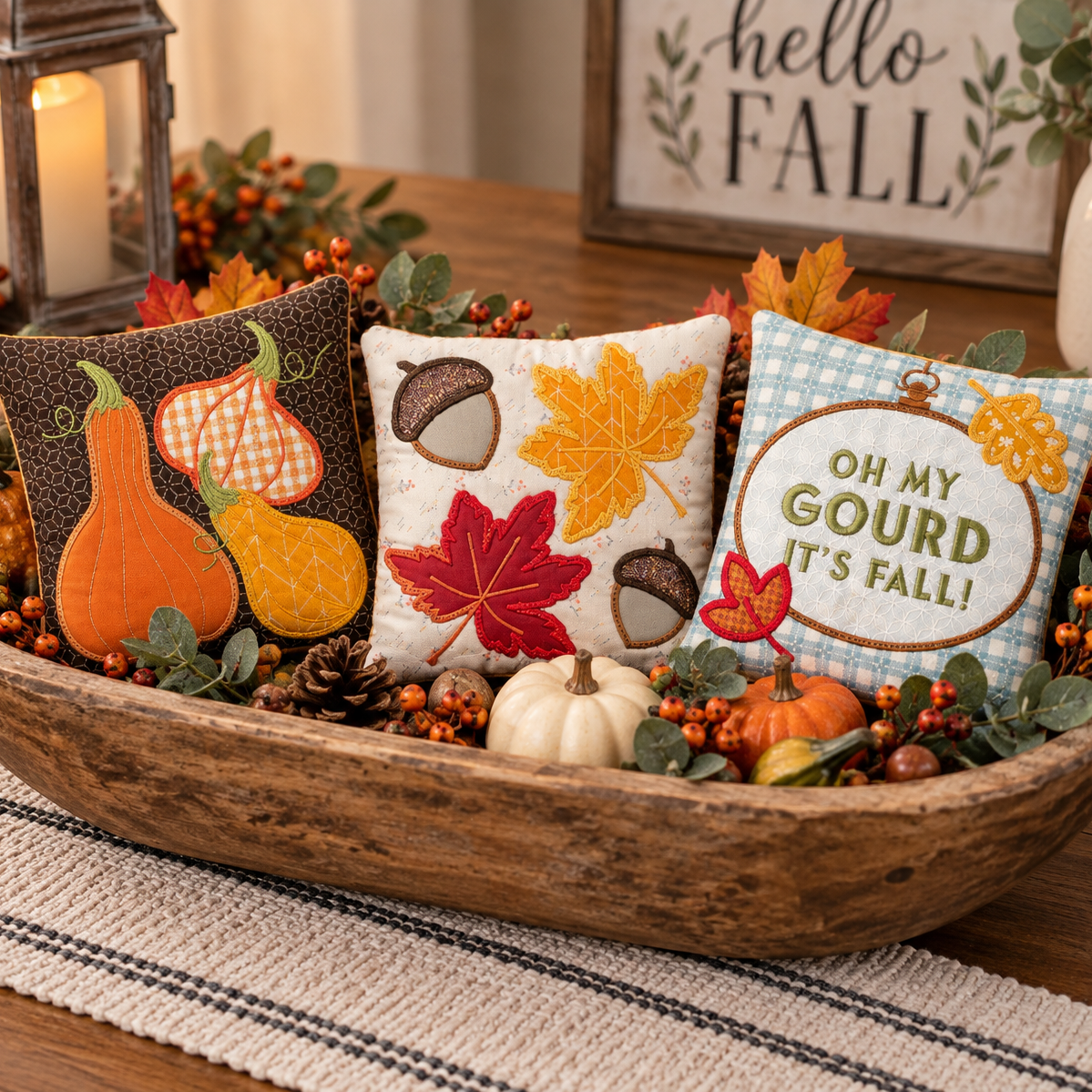 Three decorative autumn throw pillows arranged in a rustic wooden bowl centerpiece with fall leaves, berries, pinecones, and small pumpkins. The left pillow shows orange and yellow gourds on a dark brown patterned background, the center pillow features red, yellow, and brown autumn leaves and acorns on a cream background, and the right pillow reads 'OH MY GOURD IT'S FALL!' on a light blue checkered background.