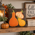 A square decorative throw pillow displayed on a wooden shelf in a fall-themed setting. The pillow features three embroidered autumn gourds in orange, yellow, and orange-and-white gingham on a dark brown geometric patterned background.