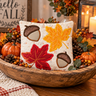 A square decorative pillow with embroidered autumn leaf and acorn motifs, styled in a rustic wooden bowl filled with fall decor including pumpkins, pinecones, and berries.