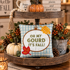 A decorative autumn-themed two-tier wooden tray centerpiece on a table. The center of the bottom tier holds a square light blue gingham pillow with the phrase “OH MY GOURD IT’S FALL!” in olive green letters, surrounded by fall leaves and berries.