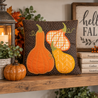 A square decorative throw pillow displayed on a wooden shelf in a fall-themed setting. The pillow features three embroidered autumn gourds in orange, yellow, and orange-and-white gingham on a dark brown geometric patterned background.