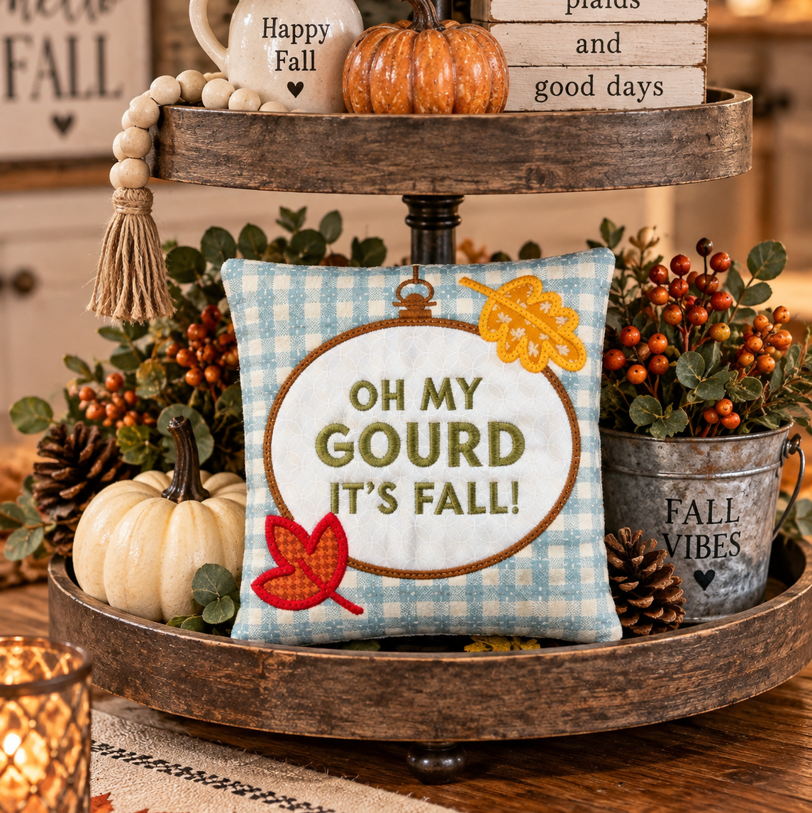 A decorative autumn-themed two-tier wooden tray centerpiece on a table. The center of the bottom tier holds a square light blue gingham pillow with the phrase “OH MY GOURD IT’S FALL!” in olive green letters, surrounded by fall leaves and berries.