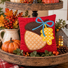 A red square decorative pillow featuring an embroidered acorn design with a blue bow and yellow leaf, displayed on a rustic wooden two-tier stand with autumn decorations.