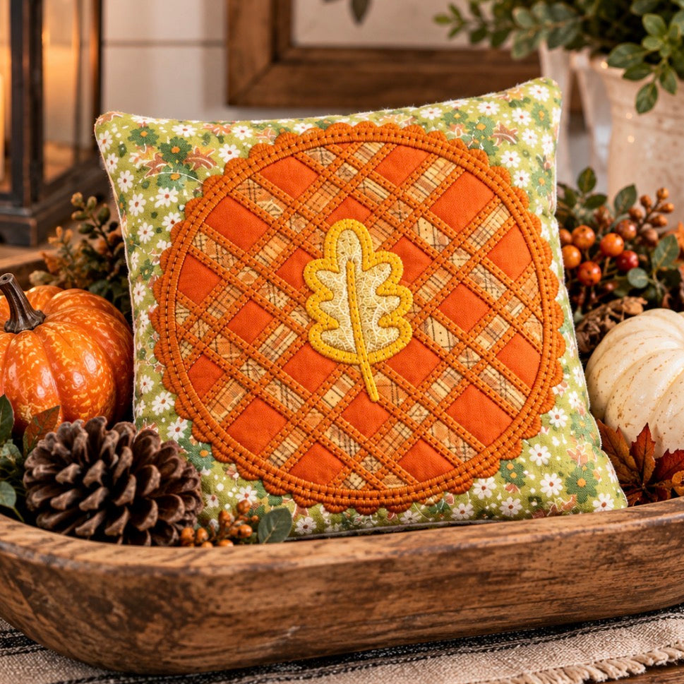 A decorative square throw pillow with an autumn theme, featuring a central orange circular patchwork design with a yellow embroidered oak leaf, set in a rustic wooden bowl with pumpkins and pinecones.
