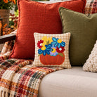 A cozy outdoor patio scene with a beige sofa styled with decorative throw pillows and a plaid fringed blanket. The central focus is a small square accent pillow featuring an embroidered orange pumpkin with colorful flowers in blue, yellow, red, and purple.