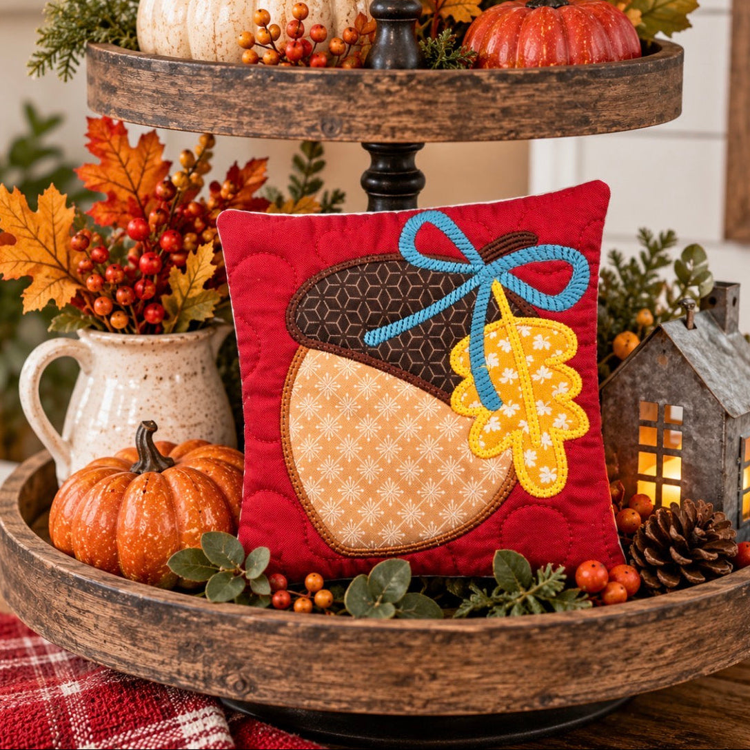 A red square decorative pillow featuring an embroidered acorn design with a blue bow and yellow leaf, displayed on a rustic wooden two-tier stand with autumn decorations.