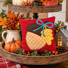 A red square decorative pillow featuring an embroidered acorn design with a blue bow and yellow leaf, displayed on a rustic wooden two-tier stand with autumn decorations.