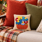 A cozy outdoor patio scene with a beige sofa styled with decorative throw pillows and a plaid fringed blanket. The central focus is a small square accent pillow featuring an embroidered orange pumpkin with colorful flowers in blue, yellow, red, and purple.