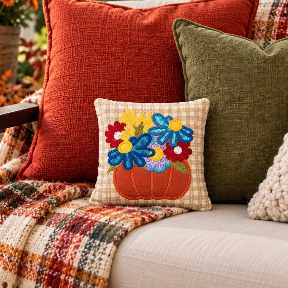 A cozy outdoor patio scene with a beige sofa styled with decorative throw pillows and a plaid fringed blanket. The central focus is a small square accent pillow featuring an embroidered orange pumpkin with colorful flowers in blue, yellow, red, and purple.