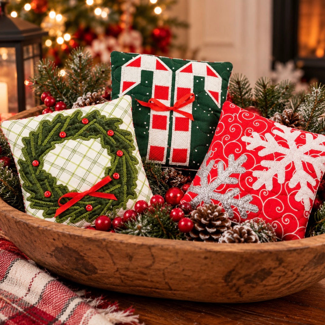 Three small Christmas-themed fabric pillows arranged in a wooden bowl with pinecones, evergreen branches, and red berries. One pillow shows a green pine wreath with red berries on a white background, one shows green and white candy canes with a red bow, and one is red with white snowflakes and a silver glittery snowflake.