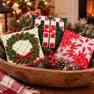 Three small Christmas-themed fabric pillows arranged in a wooden bowl with pinecones, evergreen branches, and red berries. One pillow shows a green pine wreath with red berries on a white background, one shows green and white candy canes with a red bow, and one is red with white snowflakes and a silver glittery snowflake.