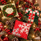 Three decorative Christmas throw pillows displayed on a decorated Christmas tree. One pillow features a green wreath design with red berries on a cream plaid background, another shows a green Christmas tree with red and white candy canes, and the third has a red background with large white snowflakes.
