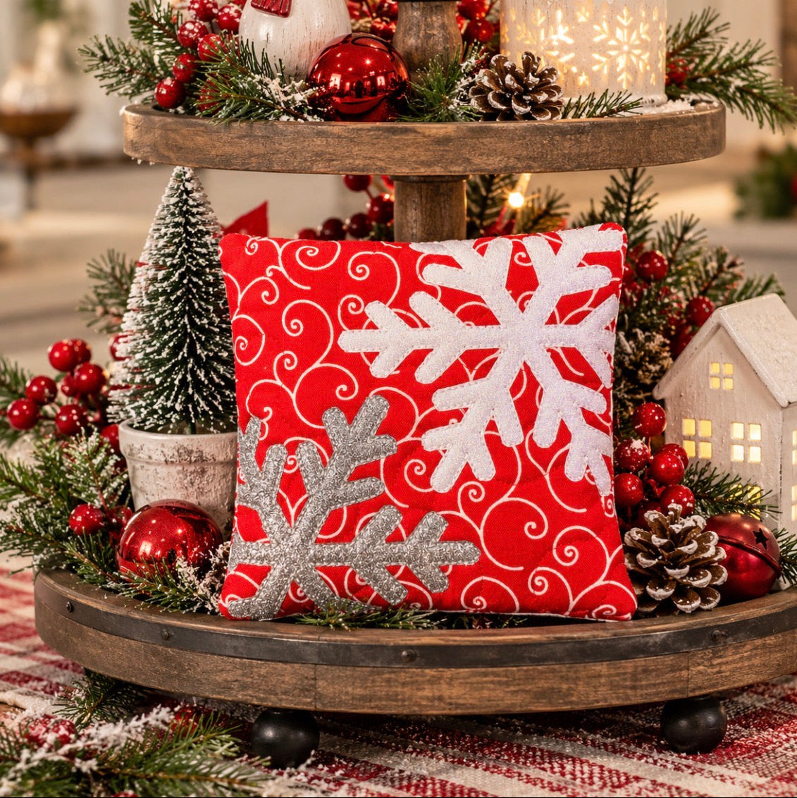 A square red Christmas throw pillow with white and silver glitter snowflakes, displayed on a rustic two-tier wooden stand surrounded by holiday greenery, ornaments, and lights.