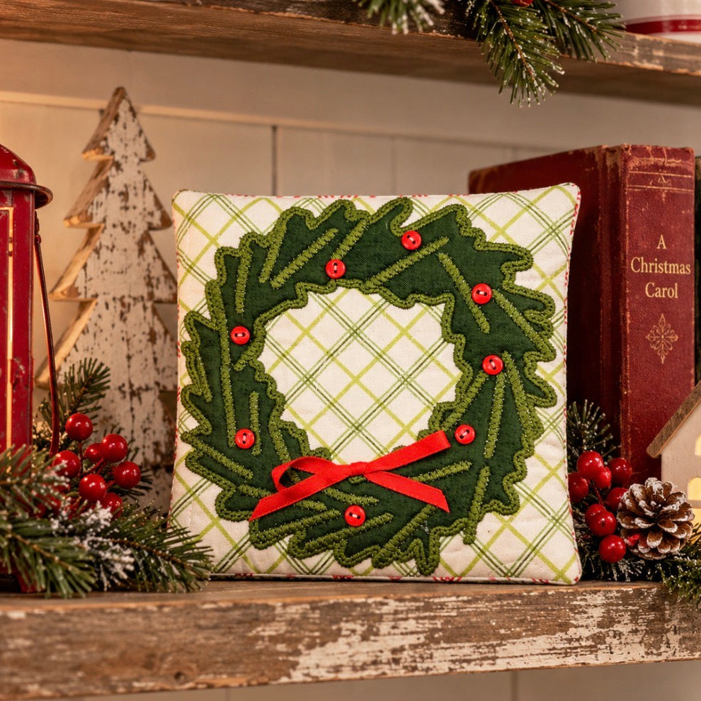 A square decorative pillow with a green holly wreath design and red ribbon bow, displayed on a rustic wooden shelf with holiday decorations, books, and a lantern.