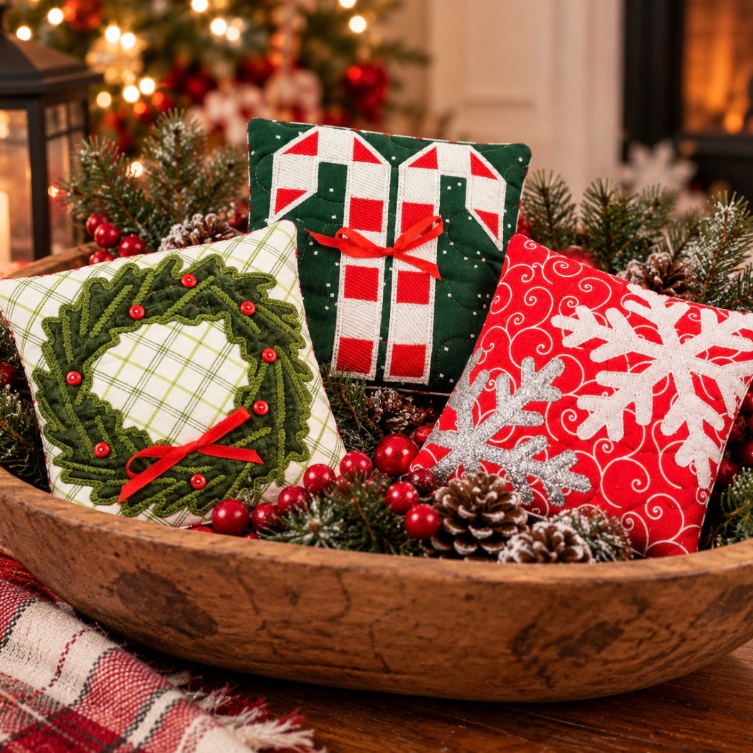 Three small Christmas-themed fabric pillows arranged in a wooden bowl with pinecones, evergreen branches, and red berries. One pillow shows a green pine wreath with red berries on a white background, one shows green and white candy canes with a red bow, and one is red with white snowflakes and a silver glittery snowflake.