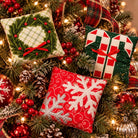 Three decorative Christmas throw pillows displayed on a decorated Christmas tree. One pillow features a green wreath design with red berries on a cream plaid background, another shows a green Christmas tree with red and white candy canes, and the third has a red background with large white snowflakes.