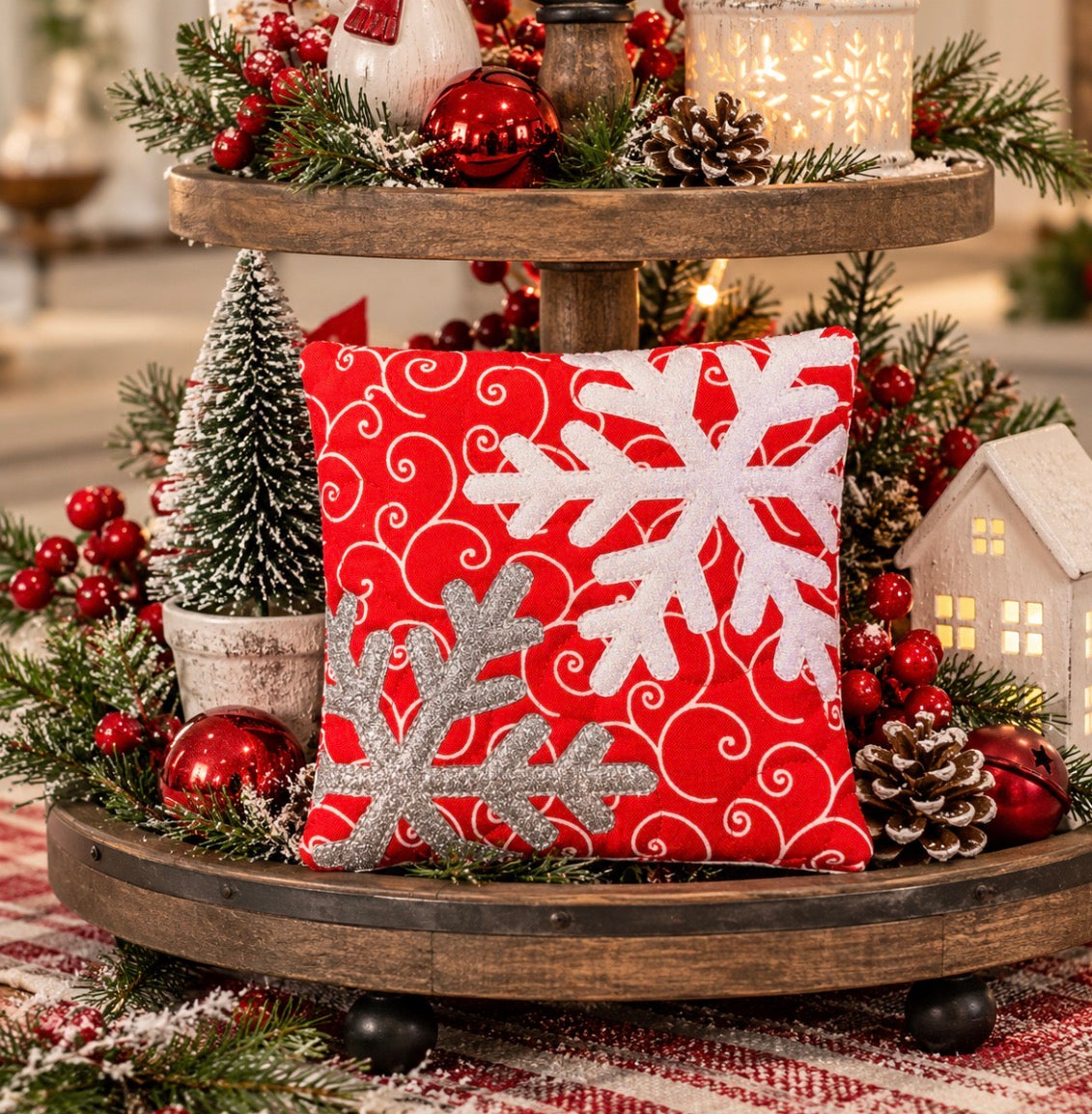 A square red Christmas throw pillow with white and silver glitter snowflakes, displayed on a rustic two-tier wooden stand surrounded by holiday greenery, ornaments, and lights.