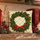 A square decorative pillow with a green holly wreath design and red ribbon bow, displayed on a rustic wooden shelf with holiday decorations, books, and a lantern.