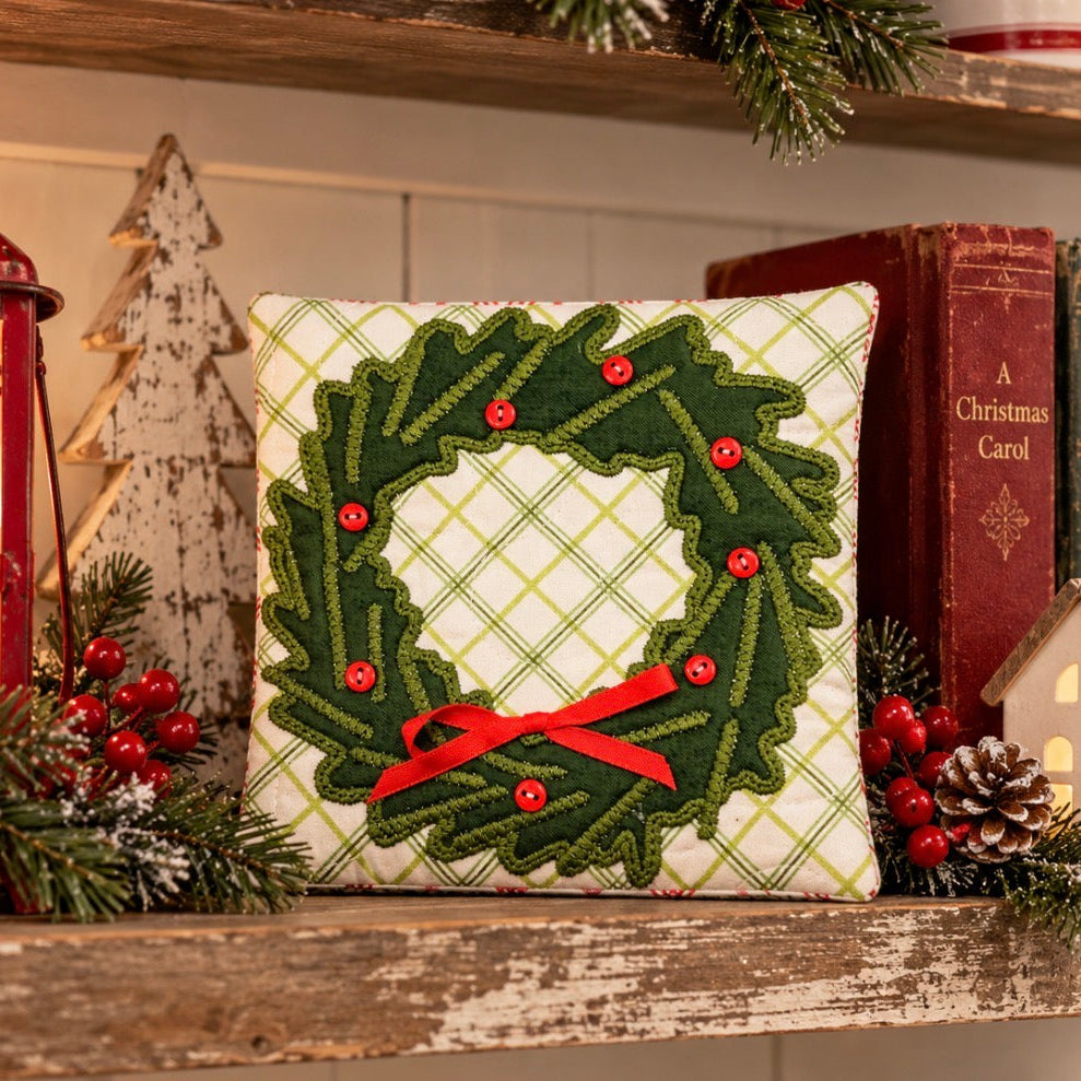 A square decorative pillow with a green holly wreath design and red ribbon bow, displayed on a rustic wooden shelf with holiday decorations, books, and a lantern.