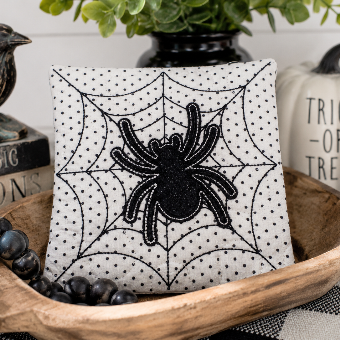 A square decorative pillow with a black spiderweb pattern and a black spider applique in the center, styled in a wooden bowl with black beads and a potted plant in the background.