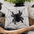 A square decorative pillow with a black spiderweb pattern and a black spider applique in the center, styled in a wooden bowl with black beads and a potted plant in the background.