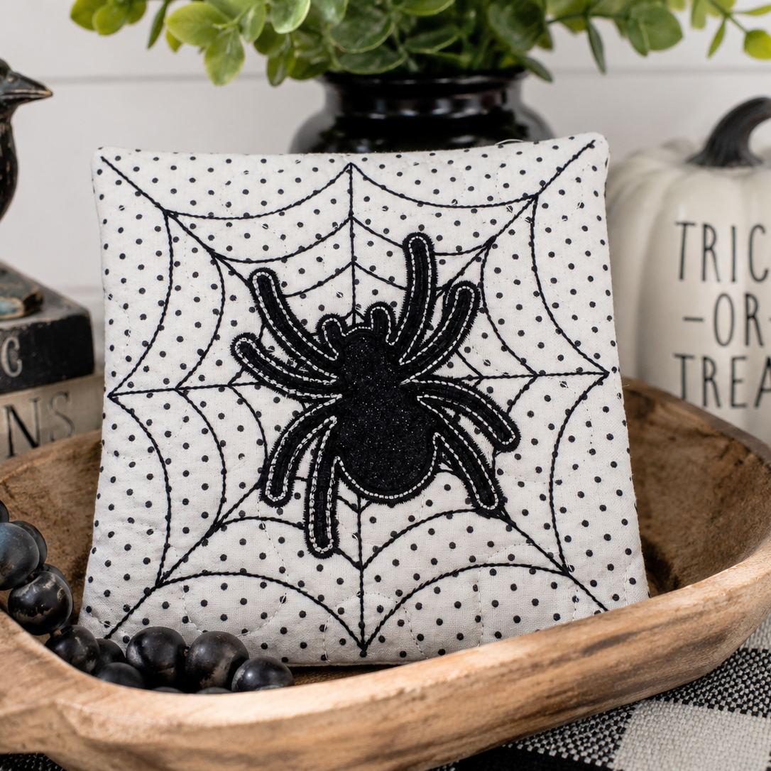 Decorative pillow with spider and web design in a bowl with Halloween-themed items.