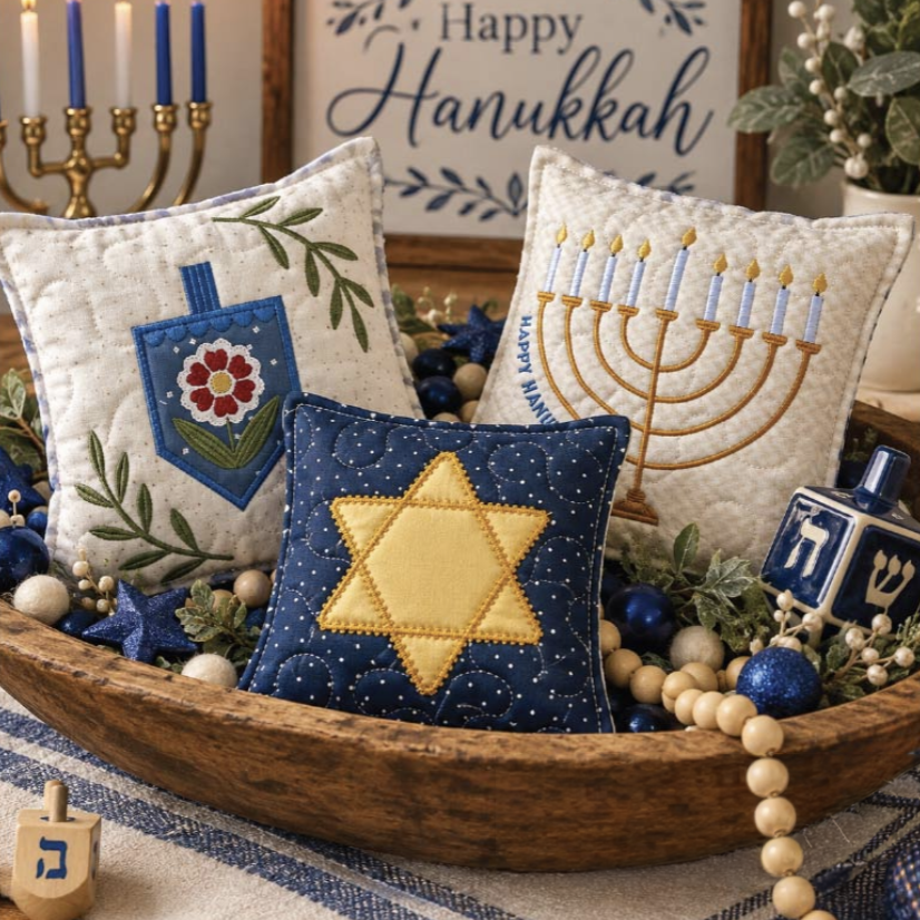 Three decorative embroidered throw pillows arranged in a wooden bowl with holiday greenery and blue ornaments. One pillow is cream with a blue Hanukkah dreidel motif, one is cream with a gold menorah, and one is navy with a yellow Star of David.