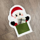 A felt Santa Claus coin purse or money pocket lies on a gray wood floor. The white felt Santa has a red hat, black and red gloves, pink cheeks, and a green money pocket with a U.