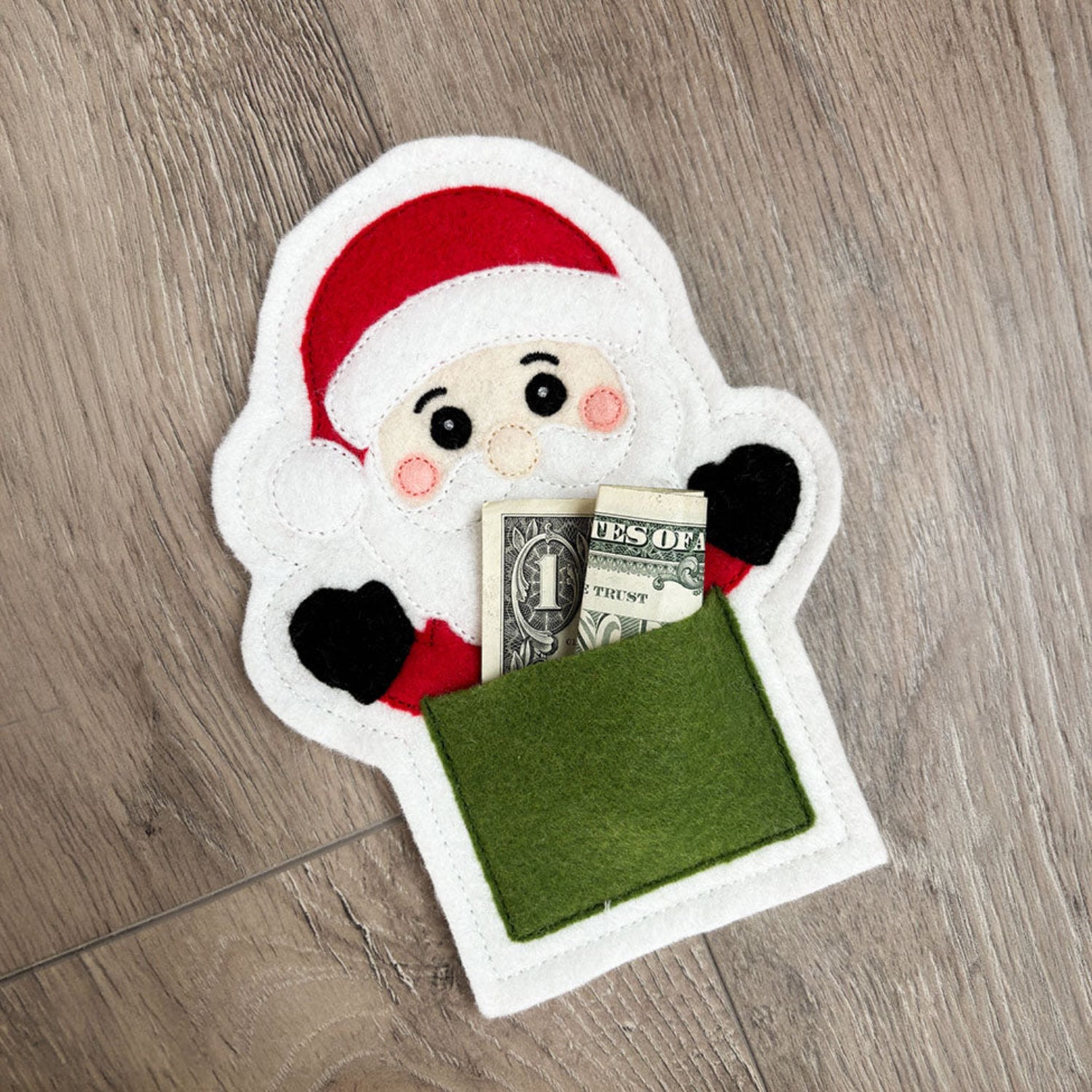 A felt Santa Claus coin purse or money pocket lies on a gray wood floor. The white felt Santa has a red hat, black and red gloves, pink cheeks, and a green money pocket with a U.