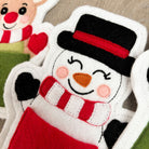 A close-up of a handmade felt Christmas stocking shaped like a snowman, featuring a black top hat with a red band, a red and white striped scarf, a black nose, and pink stitched cheeks, shown on a wooden surface.