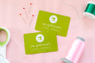 Two green gift cards with a logo and text, surrounded by sewing materials on a pink background.