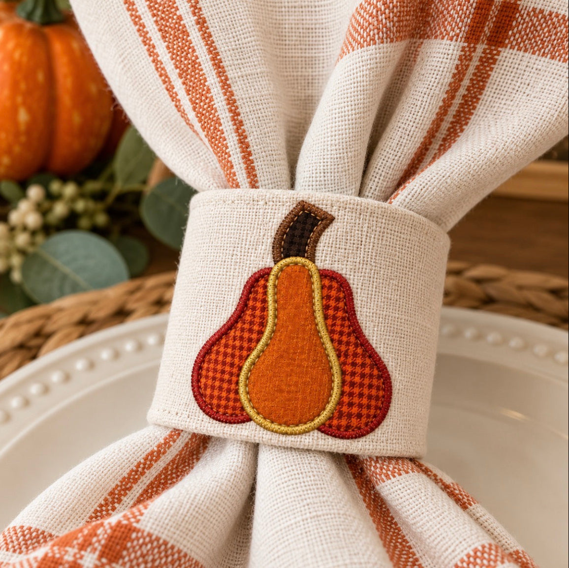 A close-up of a woven napkin with orange stripes, tied with a matching fabric napkin ring featuring an embroidered autumn pear design in orange, red, brown, and yellow.