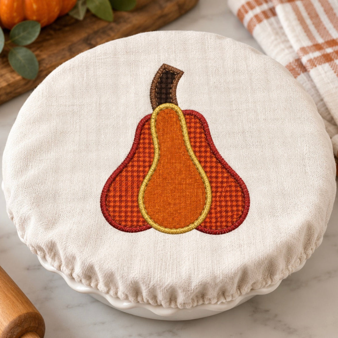 A round white fabric cover with a stitched autumn pear design in orange, brown, and yellow, shown on a white dish in a kitchen setting with a marble surface, a plaid towel, and a rolling pin.