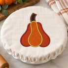 A round white fabric cover with a stitched autumn pear design in orange, brown, and yellow, shown on a white dish in a kitchen setting with a marble surface, a plaid towel, and a rolling pin.