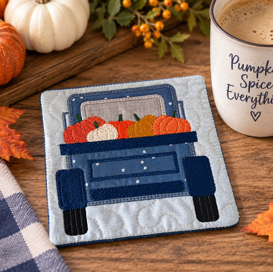 A square quilted coaster featuring an applique design of a blue pickup truck carrying orange and white pumpkins, styled on a wooden table with autumn leaves, a mug reading 'Pumpkin Spice Everything', and small pumpkins.