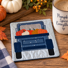 A square quilted coaster featuring an applique design of a blue pickup truck carrying orange and white pumpkins, styled on a wooden table with autumn leaves, a mug reading 'Pumpkin Spice Everything', and small pumpkins.