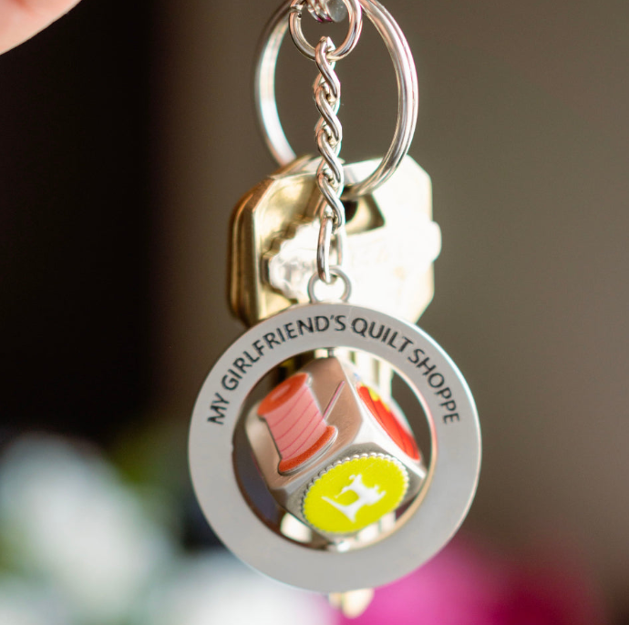 Keychain with 'My Girlfriend's Quilt Shoppe' branding on a blurred background