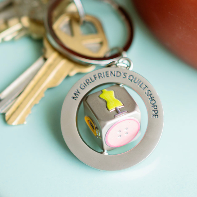 Keychain with 'My Girlfriend's Quilt Shoppe' text and quilt design on a light blue background
