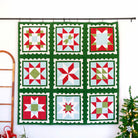 A nine-square quilted wall hanging with a green border and red, white, and light green patchwork star motifs, displayed on a black rod against a white wall, with a wooden ladder and a small decorated Christmas tree nearby.