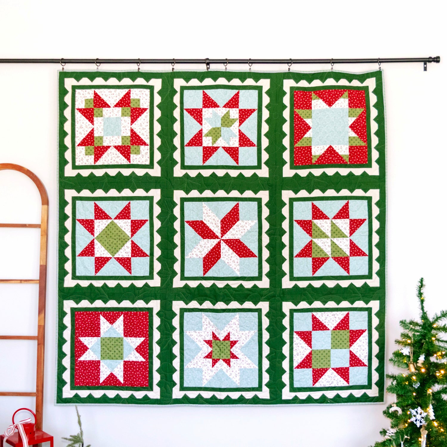 A nine-square quilted wall hanging with a green border and red, white, and light green patchwork star motifs, displayed on a black rod against a white wall, with a wooden ladder and a small decorated Christmas tree nearby.