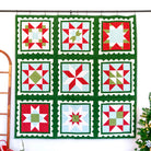 A large quilted wall hanging with a 3x3 grid of holiday starburst and snowflake-style blocks, featuring a dark green border and red, white, and light blue patterned squares, displayed on a black rod against a white wall with a wooden ladder and a small Christmas tree nearby.