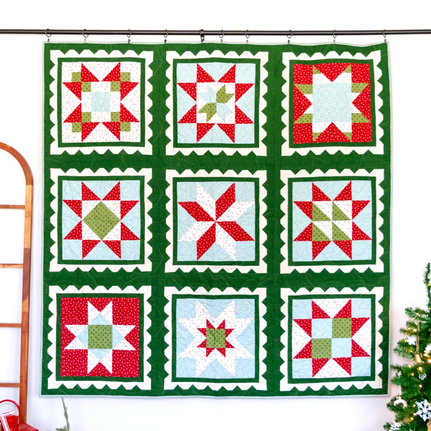 A large quilted wall hanging with a 3x3 grid of holiday starburst and snowflake-style blocks, featuring a dark green border and red, white, and light blue patterned squares, displayed on a black rod against a white wall with a wooden ladder and a small Christmas tree nearby.