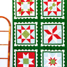 A handmade quilt with a 3x3 grid of holiday starburst patterns, featuring red, green, and white fabrics with a dark green border, hanging on a white wall next to a wooden ladder and a small Christmas tree.