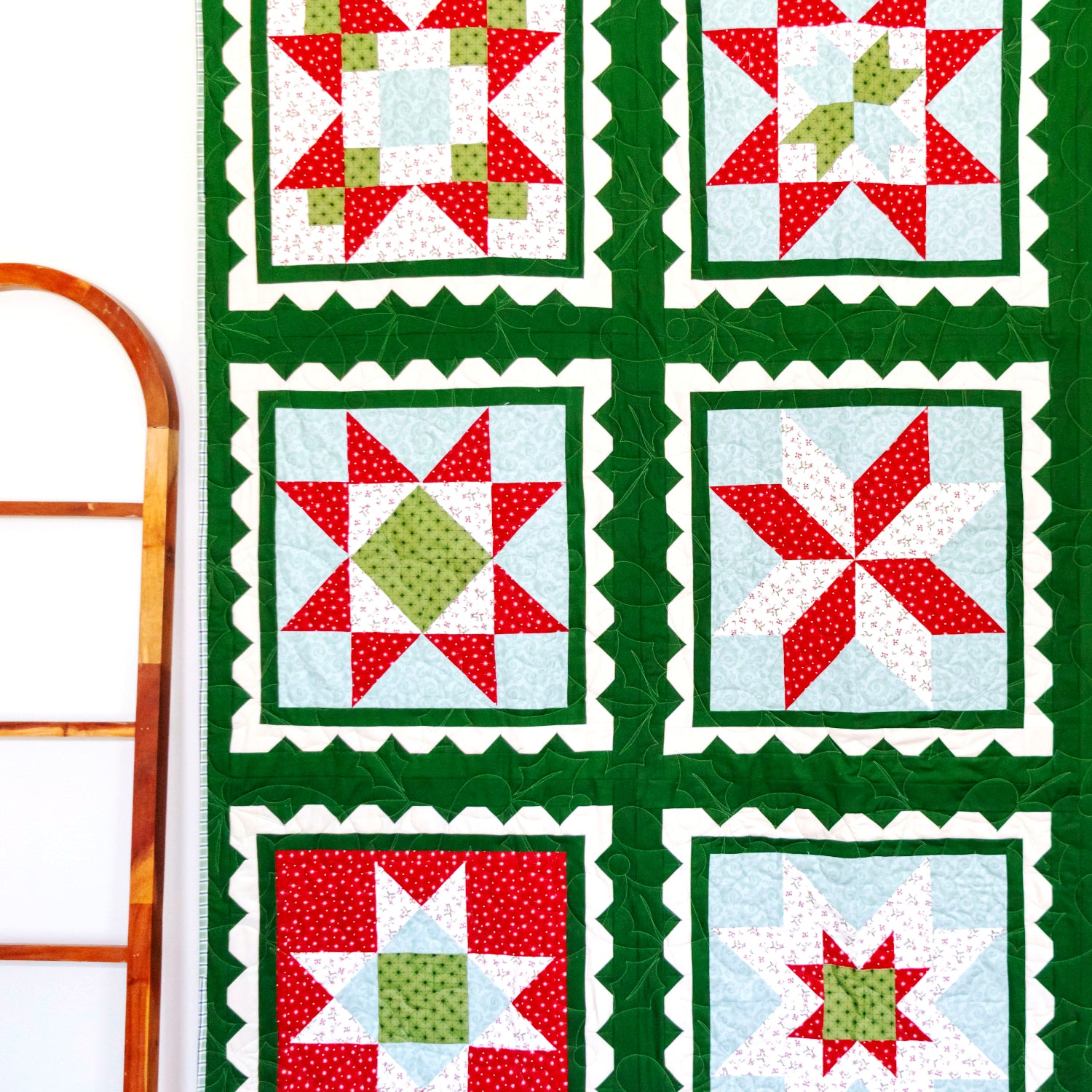 A handmade quilt with a 3x3 grid of holiday starburst patterns, featuring red, green, and white fabrics with a dark green border, hanging on a white wall next to a wooden ladder and a small Christmas tree.