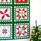 A handmade quilted wall hanging with six Christmas star and snowflake patterns in red, green, and light blue, displayed on a black metal rod against a white wall next to a decorated Christmas tree.