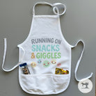 White apron with 'Running on Snacks & Giggles' text and smiley face designs on a gray background.