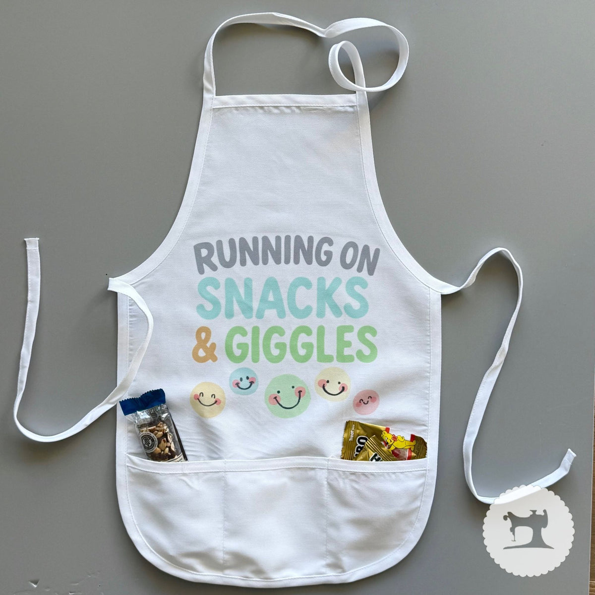 White apron with 'Running on Snacks & Giggles' text and smiley face designs on a gray background.